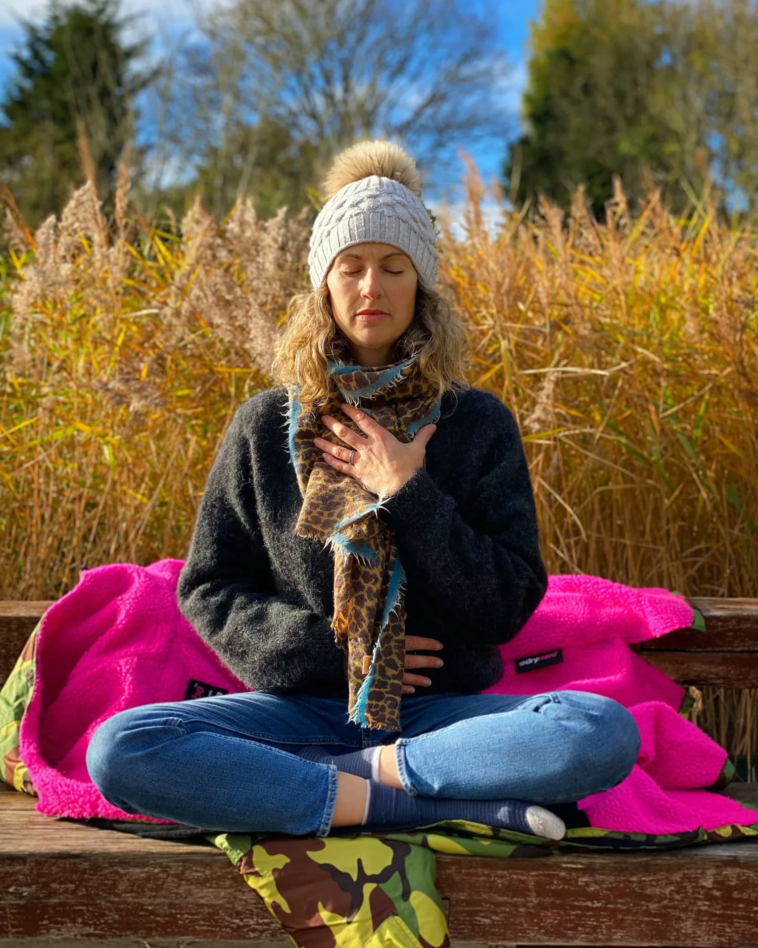 Beks meditating cross-legged on a riverside bench among golden reeds
